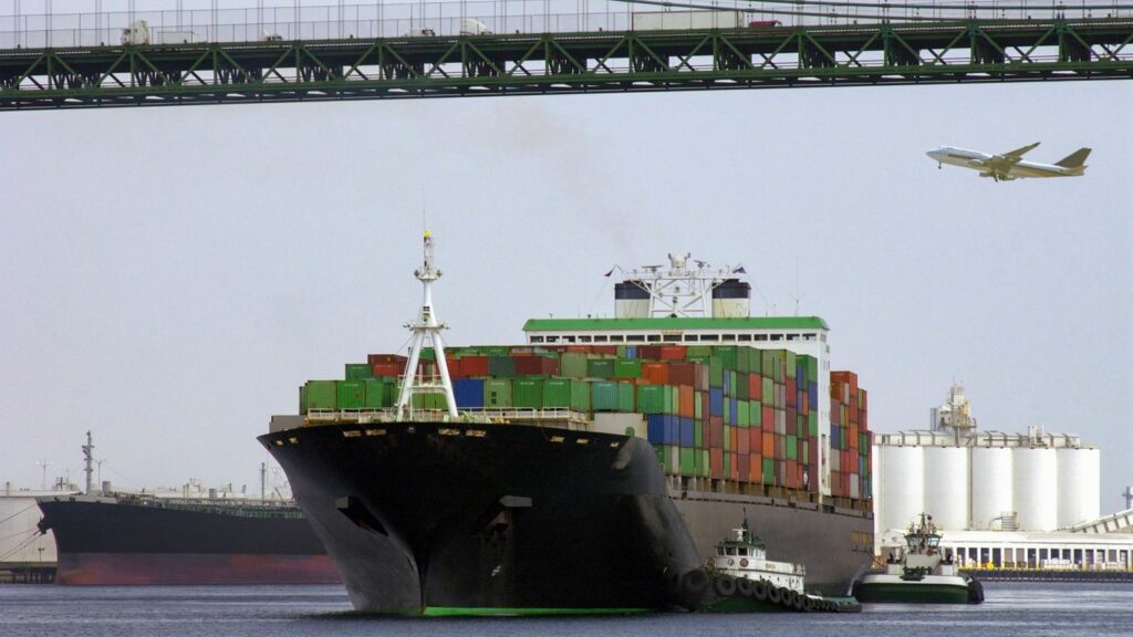 Cargo ships and tugboats pass under a bridge with an airplane overhead.