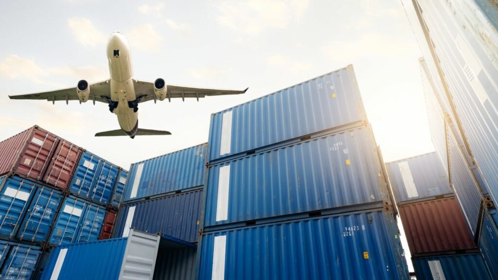Airplane flying over stacks of shipping containers under a bright sky.
