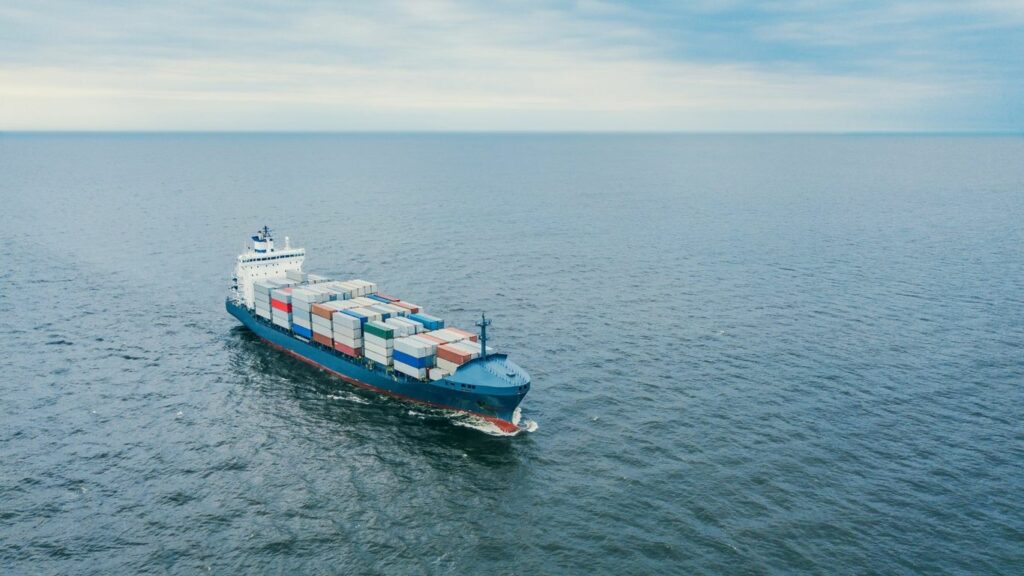 A large container ship sails across a calm, blue ocean under a cloudy sky.
